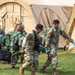 Military personnel in camouflage uniforms are carrying a patient on a litter/stretcher toward large tan military field medical tents. The scene appears to be a field medical exercise or training scenario, with personnel wearing blue medical gloves indicating a medical response context. Multiple service members are coordinating the casualty transport in an outdoor grassy area.