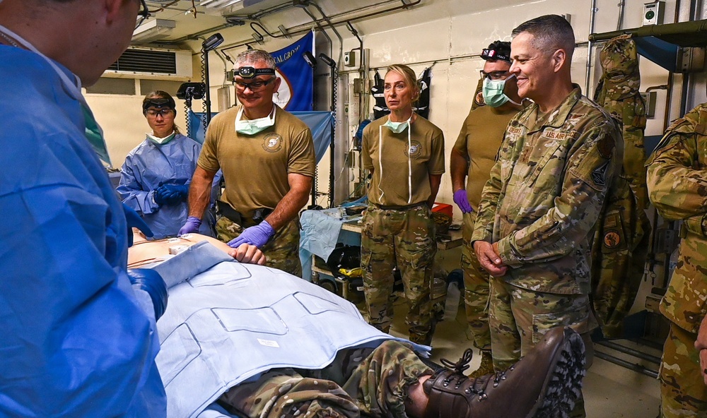 Military medical personnel in camouflage uniforms are gathered around a patient on a gurney in what appears to be a field medical facility or trauma bay. The scene depicts a medical training exercise or demonstration, with some personnel wearing surgical gloves, headlamps, and face masks. A person in blue surgical scrubs is performing or demonstrating a procedure while others observe.