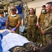 Military medical personnel in camouflage uniforms are gathered around a patient on a gurney in what appears to be a field medical facility or trauma bay. The scene depicts a medical training exercise or demonstration, with some personnel wearing surgical gloves, headlamps, and face masks. A person in blue surgical scrubs is performing or demonstrating a procedure while others observe.