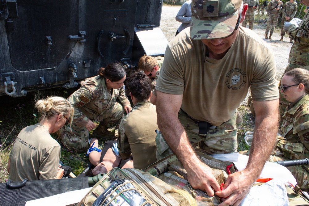 Military medical personnel are conducting a field medical training exercise near a tactical vehicle, likely a HMMWV. Multiple soldiers in OCP (Operational Camouflage Pattern) uniforms are treating a simulated casualty on the ground, with one individual accessing a medical kit on a litter. The scene appears to be an outdoor training environment with additional observers visible in the background.