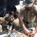 Military medical personnel are conducting a field medical training exercise near a tactical vehicle, likely a HMMWV. Multiple soldiers in OCP (Operational Camouflage Pattern) uniforms are treating a simulated casualty on the ground, with one individual accessing a medical kit on a litter. The scene appears to be an outdoor training environment with additional observers visible in the background.