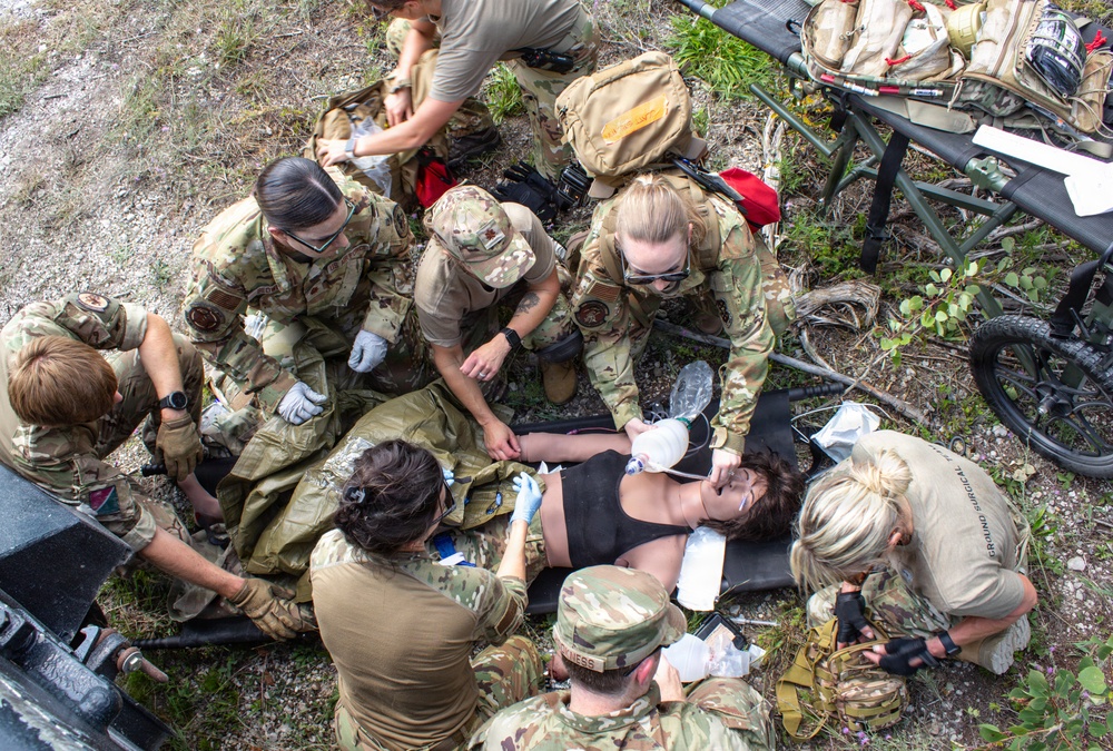 A group of military personnel in camouflage uniforms are performing tactical combat casualty care (TCCC) on a simulated patient lying on a litter/stretcher on the ground. Multiple providers are simultaneously attending to the patient, with one administering a bag-valve mask for airway management while others address other medical needs. This appears to be a training exercise based on the controlled, instructional nature of the scene.