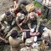 A group of military personnel in camouflage uniforms are performing tactical combat casualty care (TCCC) on a simulated patient lying on a litter/stretcher on the ground. Multiple providers are simultaneously attending to the patient, with one administering a bag-valve mask for airway management while others address other medical needs. This appears to be a training exercise based on the controlled, instructional nature of the scene.