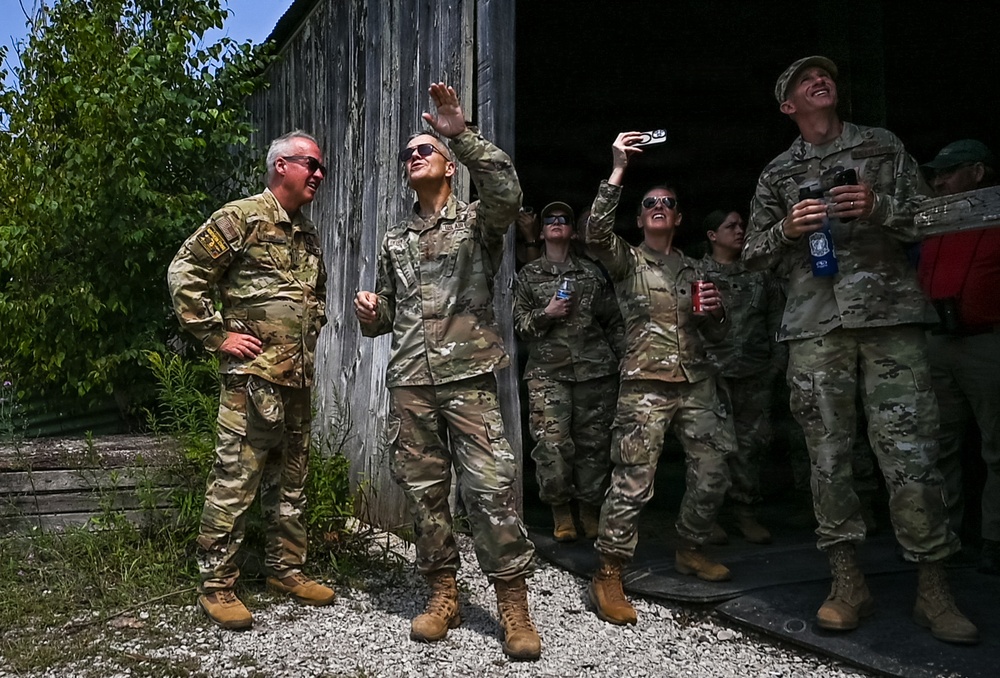 A group of military personnel in MultiCam/OCP camouflage uniforms are gathered near a weathered wooden structure, looking upward with expressions of amusement and excitement. One individual appears to be tossing or catching a small object while others observe, some holding beverages and a phone. The atmosphere appears relaxed and informal, suggesting a break or recreational moment during a training or field exercise.