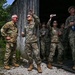 A group of military personnel in MultiCam/OCP camouflage uniforms are gathered near a weathered wooden structure, looking upward with expressions of amusement and excitement. One individual appears to be tossing or catching a small object while others observe, some holding beverages and a phone. The atmosphere appears relaxed and informal, suggesting a break or recreational moment during a training or field exercise.