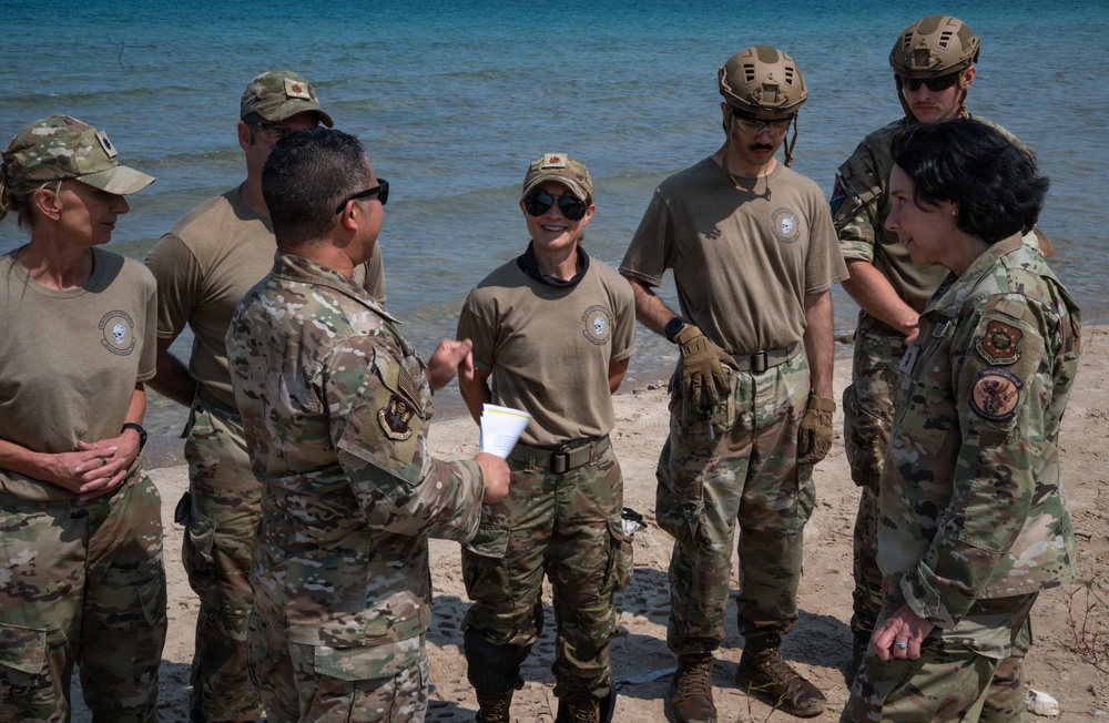 A group of approximately seven military personnel in OCP (Operational Camouflage Pattern) uniforms are gathered on a sandy beach near a body of water. One individual appears to be briefing or debriefing the group, holding papers, while others listen attentively. The group includes both male and female service members, some wearing tactical helmets and gloves.