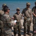 A group of approximately seven military personnel in OCP (Operational Camouflage Pattern) uniforms are gathered on a sandy beach near a body of water. One individual appears to be briefing or debriefing the group, holding papers, while others listen attentively. The group includes both male and female service members, some wearing tactical helmets and gloves.