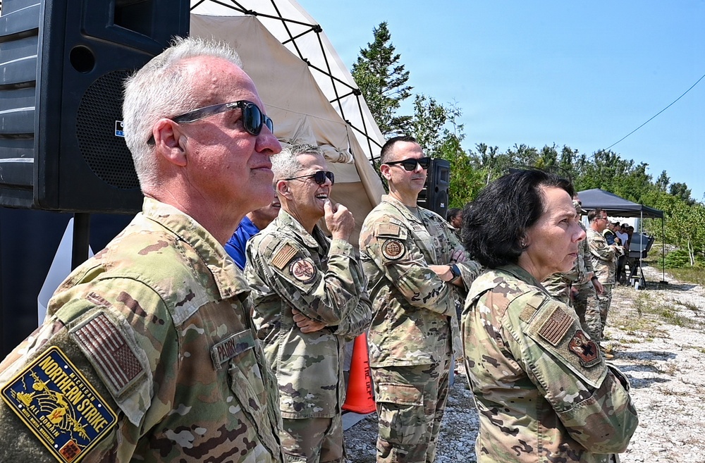 A group of U.S. military personnel in OCP (Operational Camouflage Pattern) uniforms are standing outdoors at what appears to be a field exercise or military event. The individuals are observing something off-camera, with a military tent and speaker system visible in the background. A patch on the foreground soldier's arm reads 'Northern Strike,' indicating participation in a Michigan National Guard joint training exercise.