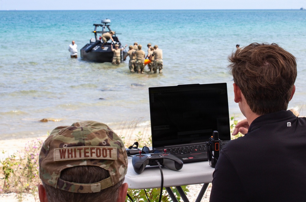 A military beach operation is underway with soldiers working around a dark tactical/unmanned surface vessel in shallow water. In the foreground, two individuals — one in military camouflage with a name tape reading 'WHITEFOOT' and one in civilian attire — monitor the operation from a folding table equipped with a laptop, a game-style controller, and a handheld radio device. The setting appears to be a training or demonstration exercise on a clear, sunny day.