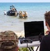 A military beach operation is underway with soldiers working around a dark tactical/unmanned surface vessel in shallow water. In the foreground, two individuals — one in military camouflage with a name tape reading 'WHITEFOOT' and one in civilian attire — monitor the operation from a folding table equipped with a laptop, a game-style controller, and a handheld radio device. The setting appears to be a training or demonstration exercise on a clear, sunny day.