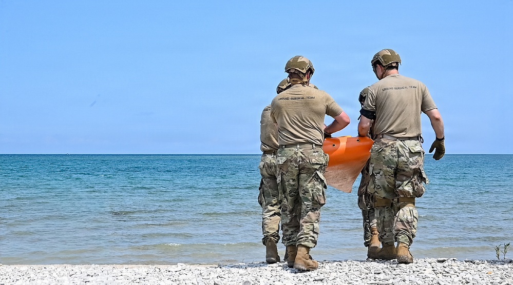 Two military personnel wearing tan/camouflage uniforms and helmets stand on a rocky shoreline facing a calm body of water. Both individuals have 'Ground Surgical Team' printed on the back of their shirts and are jointly carrying an orange object, likely a rescue or medical flotation device. The scene appears to be a training exercise or operational deployment near a large lake or sea.