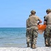 Two military personnel wearing tan/camouflage uniforms and helmets stand on a rocky shoreline facing a calm body of water. Both individuals have 'Ground Surgical Team' printed on the back of their shirts and are jointly carrying an orange object, likely a rescue or medical flotation device. The scene appears to be a training exercise or operational deployment near a large lake or sea.