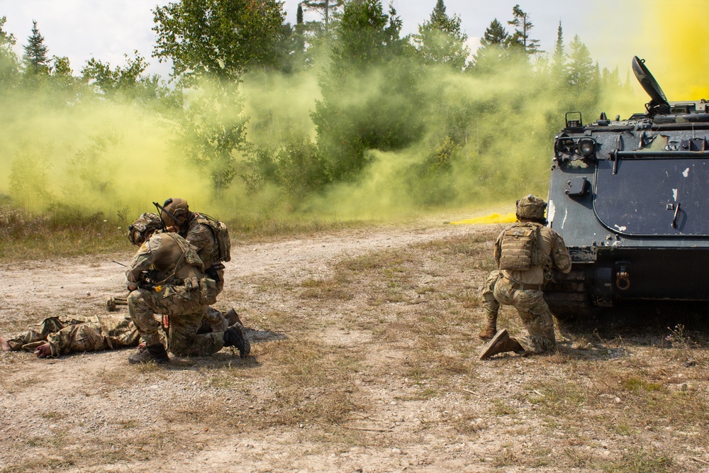 A military training exercise is underway in a wooded outdoor environment, with soldiers in full combat gear responding to a simulated casualty scenario. Yellow smoke grenades have been deployed, likely as a signal or screening measure, billowing from the direction of an armored personnel carrier on the right. Two soldiers appear to be rendering aid to a downed soldier on the ground while a third soldier kneels near the armored vehicle.