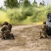 A military training exercise is underway in a wooded outdoor environment, with soldiers in full combat gear responding to a simulated casualty scenario. Yellow smoke grenades have been deployed, likely as a signal or screening measure, billowing from the direction of an armored personnel carrier on the right. Two soldiers appear to be rendering aid to a downed soldier on the ground while a third soldier kneels near the armored vehicle.