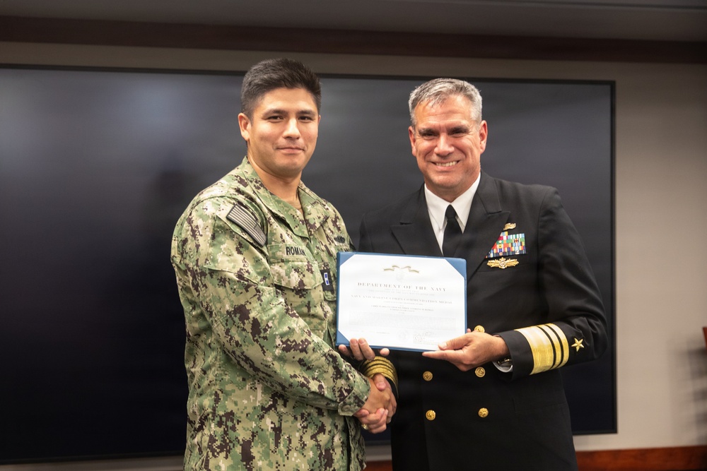 Two U.S. Navy personnel are posing together while jointly holding a Department of the Navy certificate, likely a Navy and Marine Corps Commendation Medal citation. The individual on the left is wearing NWU Type III (woodland digital camouflage) uniform with the name tape 'ROMAN' visible, while the individual on the right is wearing a Navy Service Dress Blue uniform with senior officer rank insignia and multiple service ribbons. This appears to be a formal military awards ceremony.