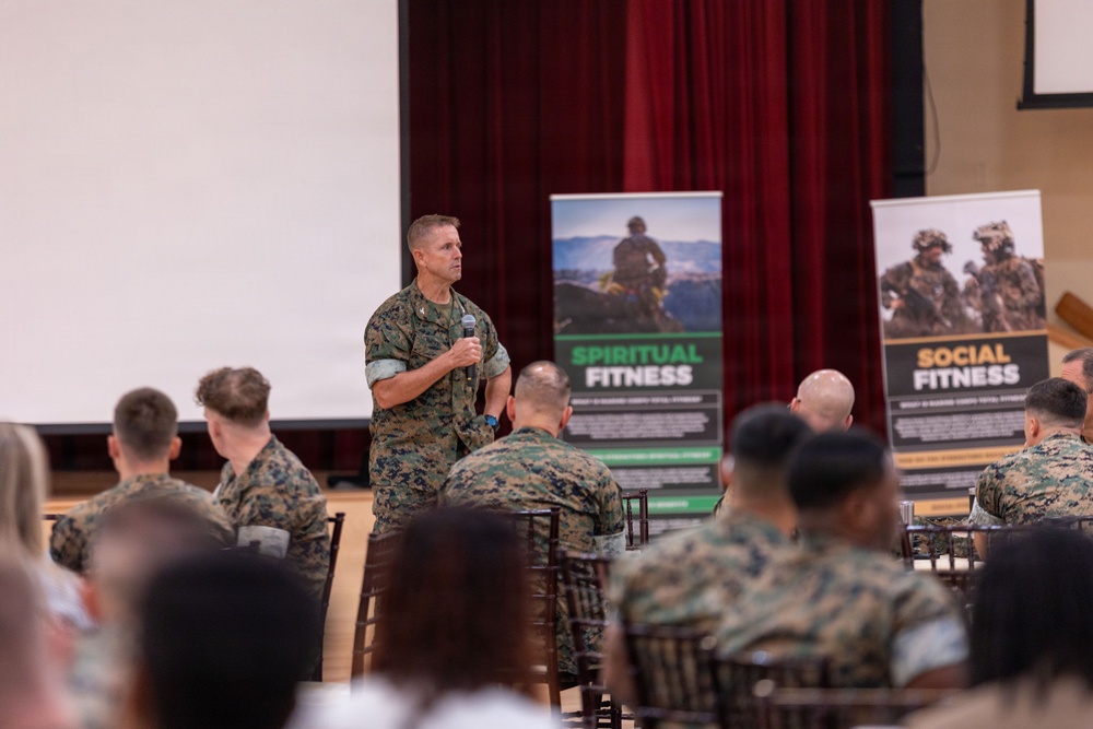 A military officer in U.S. Marine Corps camouflage uniform is addressing a seated audience of fellow Marines in what appears to be a briefing or training session. The event focuses on fitness and wellness topics, as indicated by two visible banner stands labeled 'Spiritual Fitness' and 'Social Fitness.' The setting is an auditorium or multipurpose hall with a projection screen and red curtains in the background.