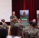 A military officer in U.S. Marine Corps camouflage uniform is addressing a seated audience of fellow Marines in what appears to be a briefing or training session. The event focuses on fitness and wellness topics, as indicated by two visible banner stands labeled 'Spiritual Fitness' and 'Social Fitness.' The setting is an auditorium or multipurpose hall with a projection screen and red curtains in the background.