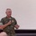 A senior military officer in U.S. Marine Corps MARPAT digital camouflage uniform is speaking into a handheld microphone, apparently addressing an audience. He is wearing gold military qualification badges on his chest and the uniform bears a 'U.S. NAVY' tape, suggesting a Navy chaplain or medical officer embedded with Marines. The setting appears to be an auditorium or briefing room with a large projection screen in the background.