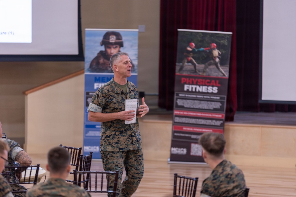 A U.S. Marine Corps officer in MARPAT camouflage uniform is addressing a group of seated Marines in what appears to be a briefing or training session. The setting is a formal hall or auditorium with projection screens and military wellness/fitness banners visible in the background. The speaker is holding papers and appears to be actively presenting or speaking to the audience.