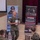 A U.S. Marine Corps officer in MARPAT camouflage uniform is addressing a group of seated Marines in what appears to be a briefing or training session. The setting is a formal hall or auditorium with projection screens and military wellness/fitness banners visible in the background. The speaker is holding papers and appears to be actively presenting or speaking to the audience.