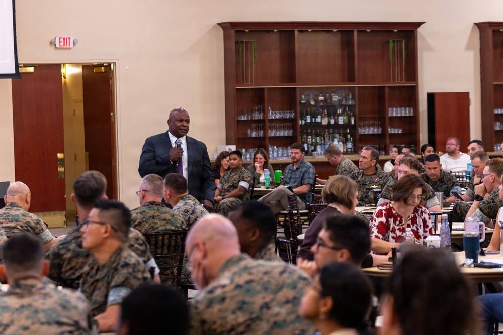 A man in a dark business suit is standing and speaking into a microphone before a large audience in what appears to be a military dining facility or event hall. The majority of the audience is wearing U.S. Marine Corps MARPAT camouflage uniforms, with some civilians also present. The setting suggests a formal briefing, training event, or guest speaker presentation.