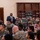 A man in a dark business suit is standing and speaking into a microphone before a large audience in what appears to be a military dining facility or event hall. The majority of the audience is wearing U.S. Marine Corps MARPAT camouflage uniforms, with some civilians also present. The setting suggests a formal briefing, training event, or guest speaker presentation.