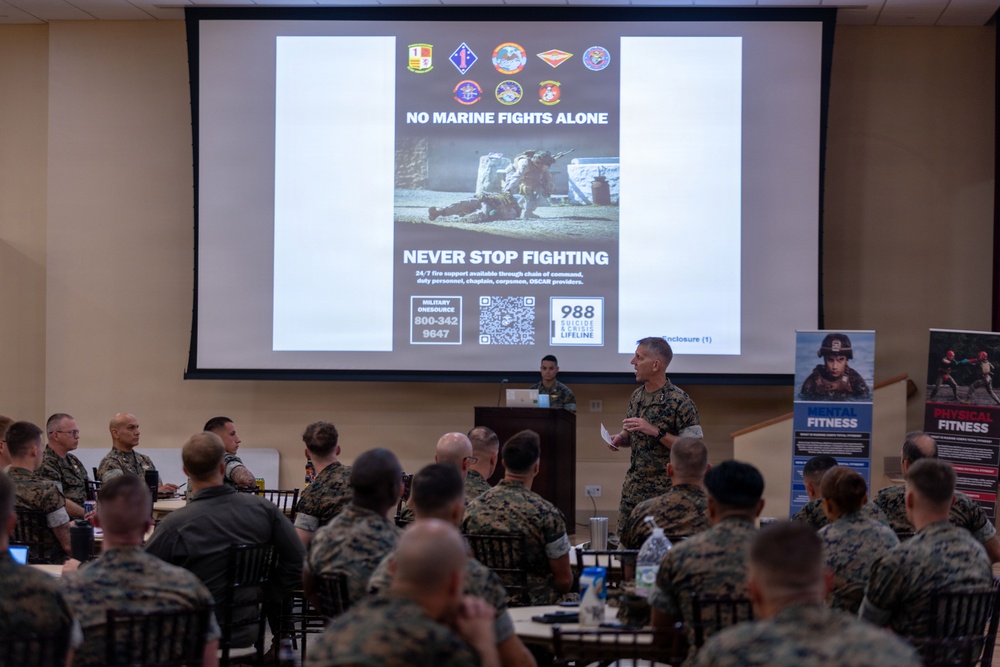 A U.S. Marine Corps briefing or training session is taking place in a conference room, with a speaker in camouflage uniform addressing a seated audience of Marines. The presentation slide displayed behind the speaker reads 'NO MARINE FIGHTS ALONE' and 'NEVER STOP FIGHTING,' indicating a mental health and suicide prevention awareness briefing. The slide includes the 988 Suicide and Crisis Lifeline number and Military OneSource contact information (800-342-9647).