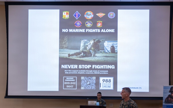 A U.S. Marine Corps briefing or training session is taking place in a conference room, with a speaker in camouflage uniform addressing a seated audience of Marines. The presentation slide displayed behind the speaker reads 'NO MARINE FIGHTS ALONE' and 'NEVER STOP FIGHTING,' indicating a mental health and suicide prevention awareness briefing. The slide includes the 988 Suicide and Crisis Lifeline number and Military OneSource contact information (800-342-9647).