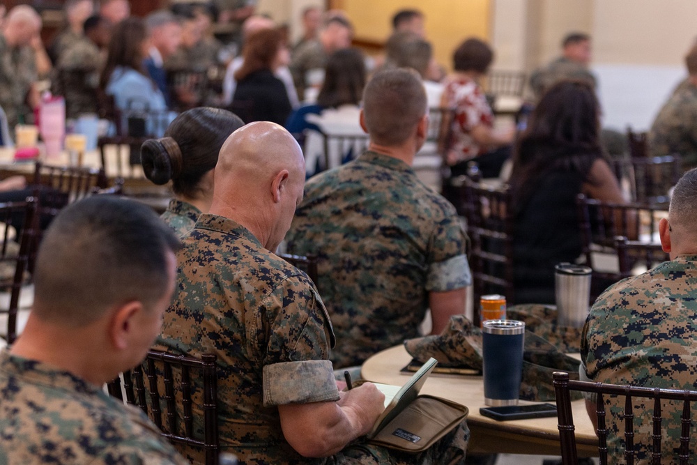 A group of military personnel, wearing MARPAT (Marine Corps digital camouflage) uniforms, are seated at tables in what appears to be a briefing, training session, or formal gathering. The foreground shows several Marines from behind, with one taking notes in a notebook or tablet folio. The setting appears to be a large hall or event space with multiple tables and a mix of uniformed and civilian attendees.