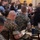 A group of military personnel, wearing MARPAT (Marine Corps digital camouflage) uniforms, are seated at tables in what appears to be a briefing, training session, or formal gathering. The foreground shows several Marines from behind, with one taking notes in a notebook or tablet folio. The setting appears to be a large hall or event space with multiple tables and a mix of uniformed and civilian attendees.
