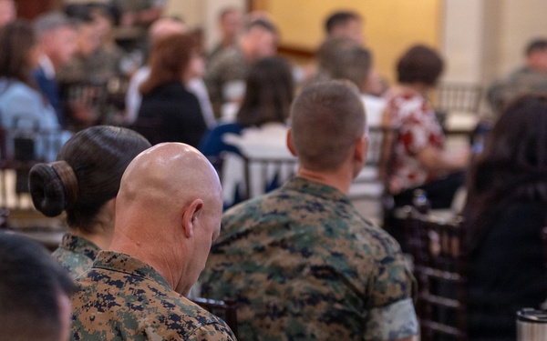 A group of military personnel, wearing MARPAT (Marine Corps digital camouflage) uniforms, are seated at tables in what appears to be a briefing, training session, or formal gathering. The foreground shows several Marines from behind, with one taking notes in a notebook or tablet folio. The setting appears to be a large hall or event space with multiple tables and a mix of uniformed and civilian attendees.