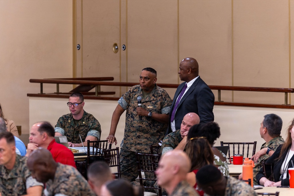 A military briefing or conference is taking place in a large hall, with a mix of uniformed military personnel and civilians seated at tables. A Marine in camouflage uniform is standing and speaking into a microphone, accompanied by a civilian in a dark suit with a purple tie. The setting appears to be a formal military event or town hall meeting.