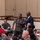 A military briefing or conference is taking place in a large hall, with a mix of uniformed military personnel and civilians seated at tables. A Marine in camouflage uniform is standing and speaking into a microphone, accompanied by a civilian in a dark suit with a purple tie. The setting appears to be a formal military event or town hall meeting.
