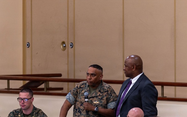 A military briefing or conference is taking place in a large hall, with a mix of uniformed military personnel and civilians seated at tables. A Marine in camouflage uniform is standing and speaking into a microphone, accompanied by a civilian in a dark suit with a purple tie. The setting appears to be a formal military event or town hall meeting.