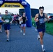 A running race is underway at Naval Air Station Cape Canaveral, organized by MWR (Morale, Welfare and Recreation). Participants of various ages and fitness levels are passing through an inflatable START/FINISH arch on a paved path. The event appears to be a 5K or fun run held in the early morning based on the overcast lighting conditions.