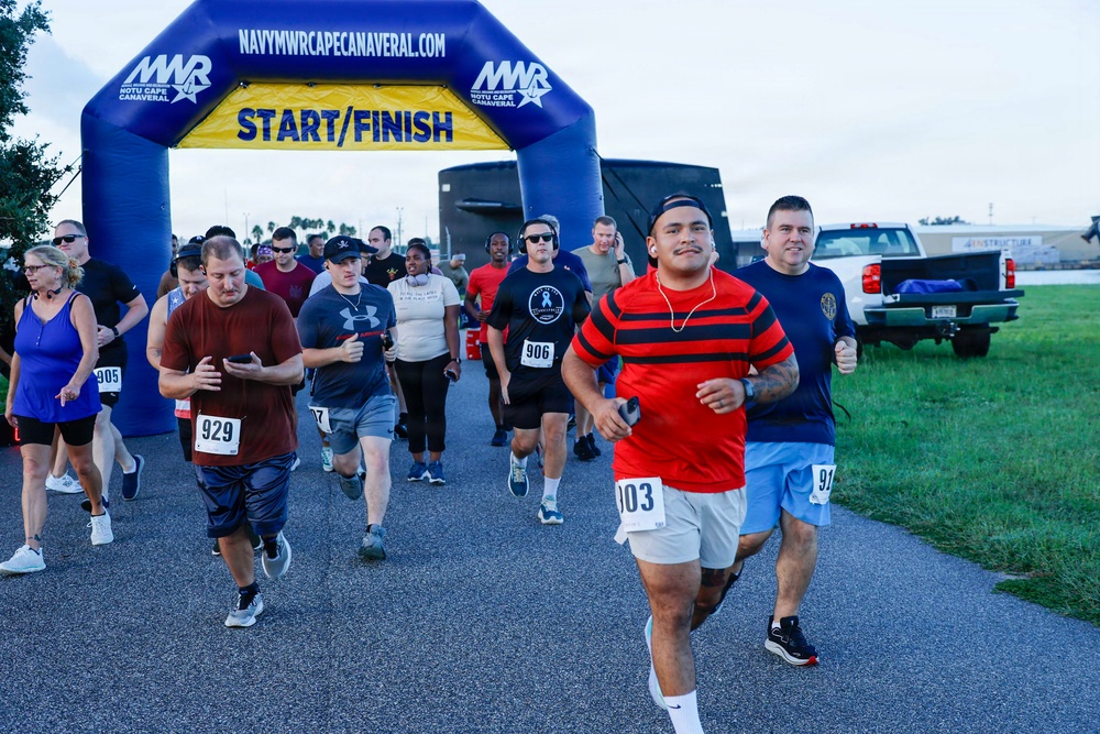 A group of runners are crossing or departing from a START/FINISH arch at a road race organized by Navy MWR Cape Canaveral. Participants wearing numbered bibs are running on a paved path in what appears to be an outdoor military installation setting. The event appears to be a community fun run or 5K race held during overcast conditions.