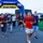 A group of runners are crossing or departing from a START/FINISH arch at a road race organized by Navy MWR Cape Canaveral. Participants wearing numbered bibs are running on a paved path in what appears to be an outdoor military installation setting. The event appears to be a community fun run or 5K race held during overcast conditions.
