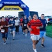 A group of runners are crossing or departing from a START/FINISH arch at a road race organized by Navy MWR Cape Canaveral. Participants wearing numbered bibs are running on a paved path in what appears to be an outdoor military installation setting. The event appears to be a community fun run or 5K race held during overcast conditions.