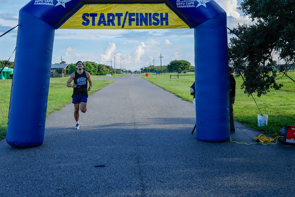 A male runner is approaching the Start/Finish inflatable arch at a road race event at NOTU Cape Canaveral. The runner is wearing a Magic basketball jersey and blue shorts, appearing to be near the finish line. An event official or volunteer stands to the right side of the arch monitoring the finish area.