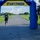 A male runner is approaching the Start/Finish inflatable arch at a road race event at NOTU Cape Canaveral. The runner is wearing a Magic basketball jersey and blue shorts, appearing to be near the finish line. An event official or volunteer stands to the right side of the arch monitoring the finish area.