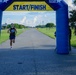 A male runner is approaching the Start/Finish inflatable arch at a road race event at NOTU Cape Canaveral. The runner is wearing a Magic basketball jersey and blue shorts, appearing to be near the finish line. An event official or volunteer stands to the right side of the arch monitoring the finish area.