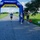 A running race event at Navy MWR Cape Canaveral, featuring a blue and yellow inflatable START/FINISH arch. A male runner wearing bib number 928 is approaching the finish line on a paved path, while two event staff or volunteers stand near the arch. The setting is an open grassy field under a partly cloudy sky.