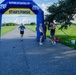 A running race event at Navy MWR Cape Canaveral, featuring a blue and yellow inflatable START/FINISH arch. A male runner wearing bib number 928 is approaching the finish line on a paved path, while two event staff or volunteers stand near the arch. The setting is an open grassy field under a partly cloudy sky.