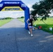 A runner wearing bib number 408 crosses through an inflatable START/FINISH arch at a road race organized by Navy MWR Cape Canaveral. The event takes place on a paved road surrounded by green grass fields under a partly cloudy sky. The runner is dressed in a black tank top, white shorts, white sneakers, and sunglasses.