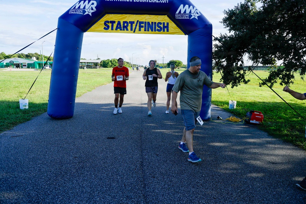 A road race event at NOTU Cape Canaveral, organized by MWR (Morale, Welfare and Recreation), showing runners approaching the Start/Finish inflatable arch. Several participants wearing race bibs are visible mid-race on a paved path surrounded by green grass. A race official or volunteer is positioned near the finish line area.