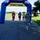 A road race event at NOTU Cape Canaveral, organized by MWR (Morale, Welfare and Recreation), showing runners approaching the Start/Finish inflatable arch. Several participants wearing race bibs are visible mid-race on a paved path surrounded by green grass. A race official or volunteer is positioned near the finish line area.