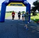 A road race event at NOTU Cape Canaveral, organized by MWR (Morale, Welfare and Recreation), showing runners approaching the Start/Finish inflatable arch. Several participants wearing race bibs are visible mid-race on a paved path surrounded by green grass. A race official or volunteer is positioned near the finish line area.