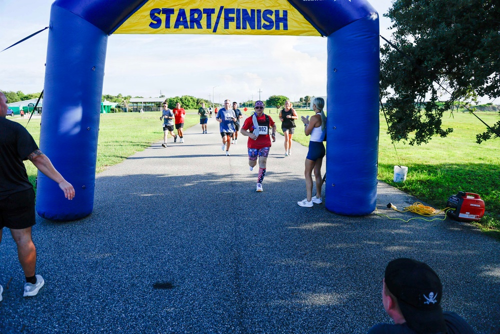 A group of runners approaches a blue and yellow inflatable START/FINISH arch during an outdoor road race. The lead runner, wearing bib number 902 and patriotic-themed attire including American flag leggings and red/white/blue accessories, is nearest to the finish line. Spectators and race officials line the sides of the path cheering the participants on.