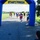 A group of runners approaches a blue and yellow inflatable START/FINISH arch during an outdoor road race. The lead runner, wearing bib number 902 and patriotic-themed attire including American flag leggings and red/white/blue accessories, is nearest to the finish line. Spectators and race officials line the sides of the path cheering the participants on.