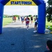 A group of runners approaches a blue and yellow inflatable START/FINISH arch during an outdoor road race. The lead runner, wearing bib number 902 and patriotic-themed attire including American flag leggings and red/white/blue accessories, is nearest to the finish line. Spectators and race officials line the sides of the path cheering the participants on.